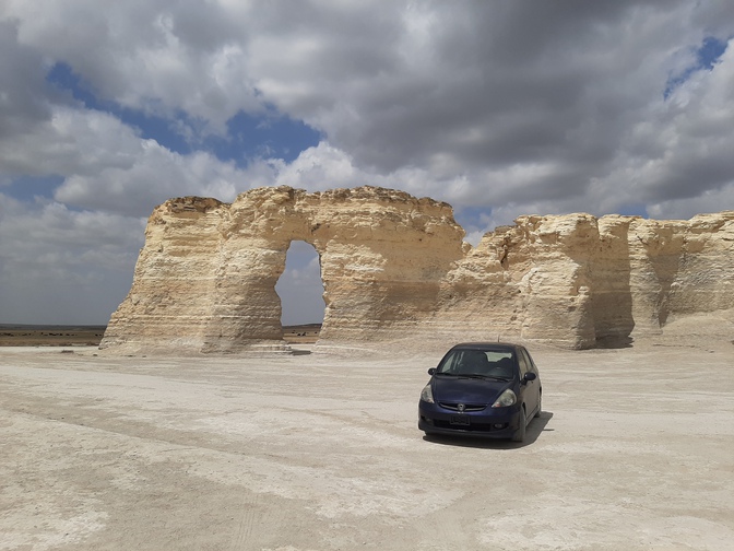 A purple Honda Fit parked serenely in front of a large chalk rock formation called 'Monument Rocks' in Kansas, under billowing clouds, miles from anything and anyone else.
